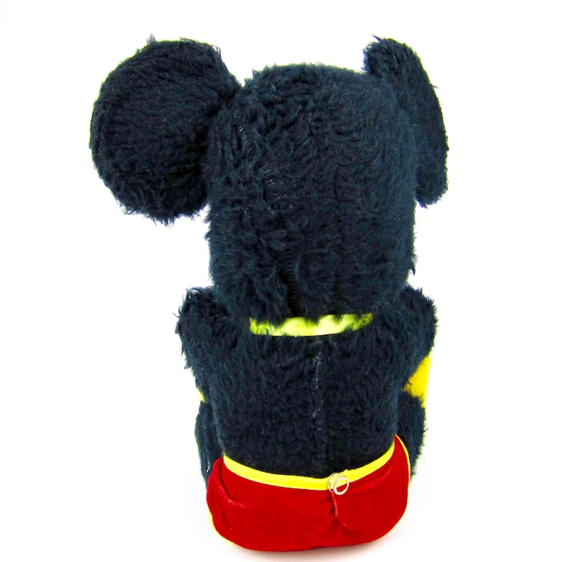 Puede incluir: Juguete de peluche negro de Mickey Mouse sentado de espaldas a la c&aacute;mara. El juguete lleva pantalones cortos rojos con un cintur&oacute;n amarillo y tiene grandes orejas negras.