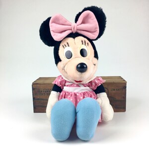 May include: A plush Minnie Mouse toy wearing a pink dress with white polka dots, a pink bow, and blue shoes. The toy is sitting on a wooden crate.