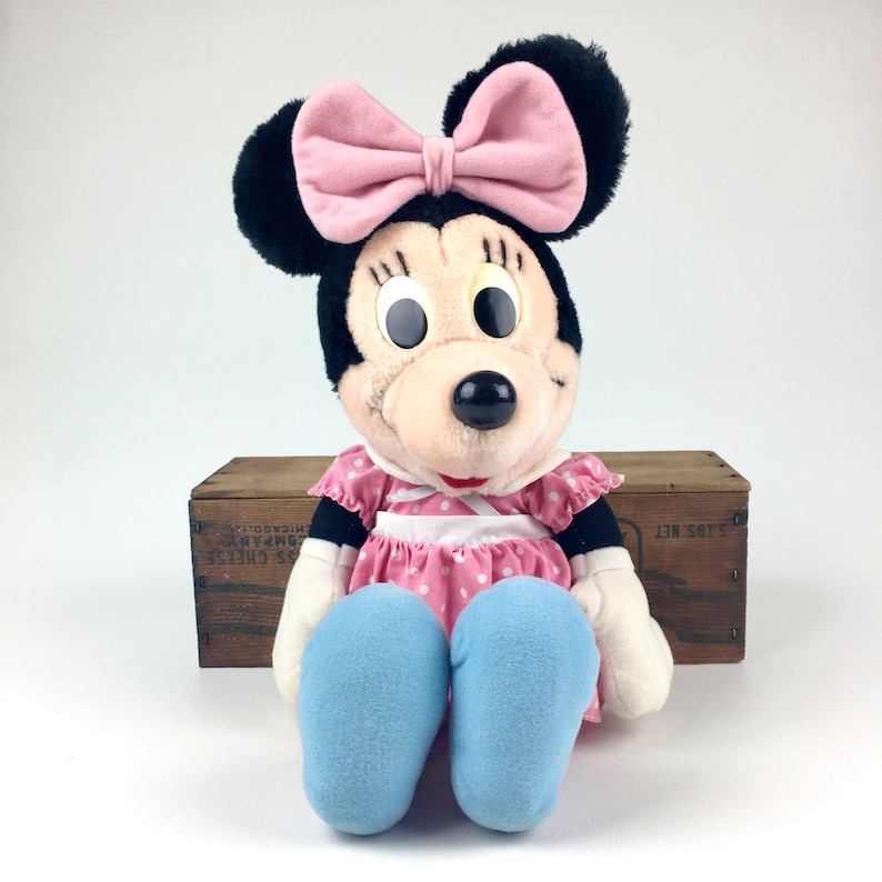 May include: A plush Minnie Mouse toy wearing a pink dress with white polka dots, a pink bow, and blue shoes. The toy is sitting on a wooden crate.