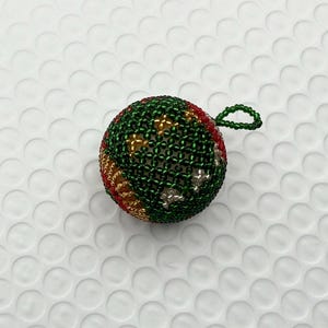 May include: A beaded Christmas ornament. The ornament is a sphere, primarily green with red, gold, and silver accents. It has a small loop at the top for hanging. The beads are tightly woven, creating a textured surface.