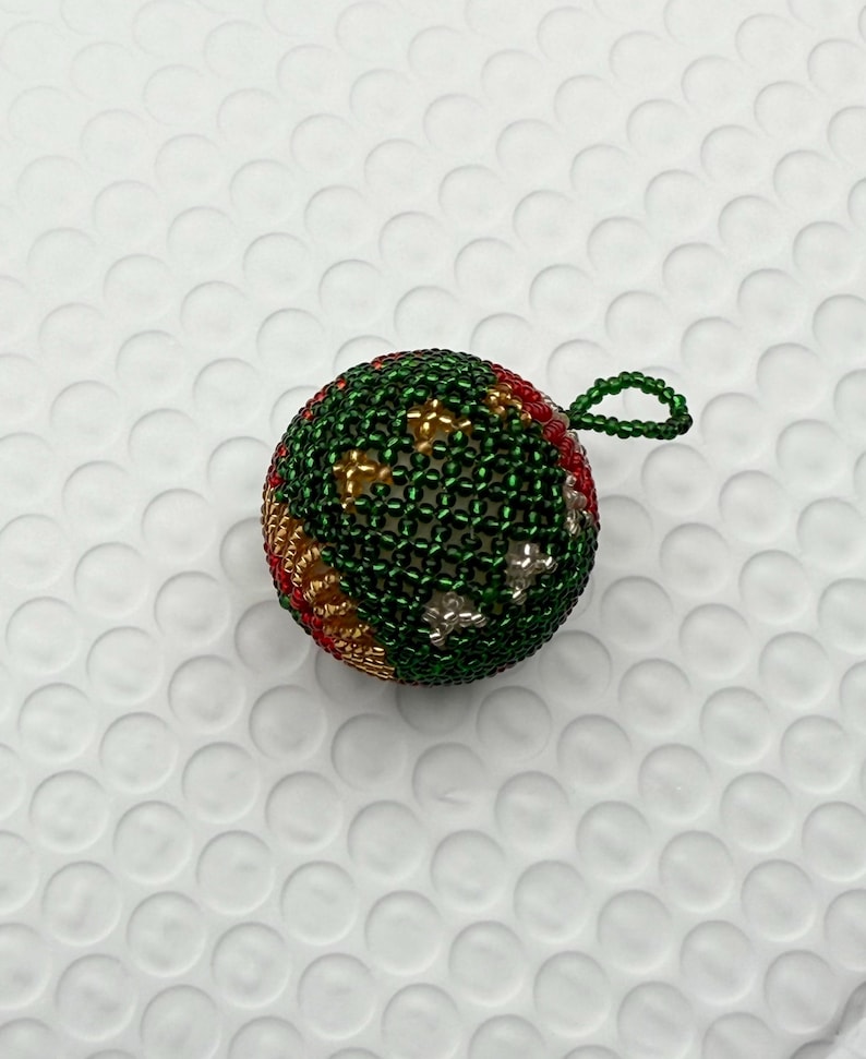 May include: A beaded Christmas ornament. The ornament is a sphere, primarily green with red, gold, and silver accents. It has a small loop at the top for hanging. The beads are tightly woven, creating a textured surface.