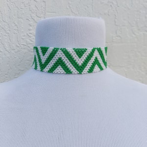 May include: A green and white beaded choker necklace with a chevron pattern. The necklace is displayed on a white mannequin head. The choker is made of small, round beads and has a geometric design.