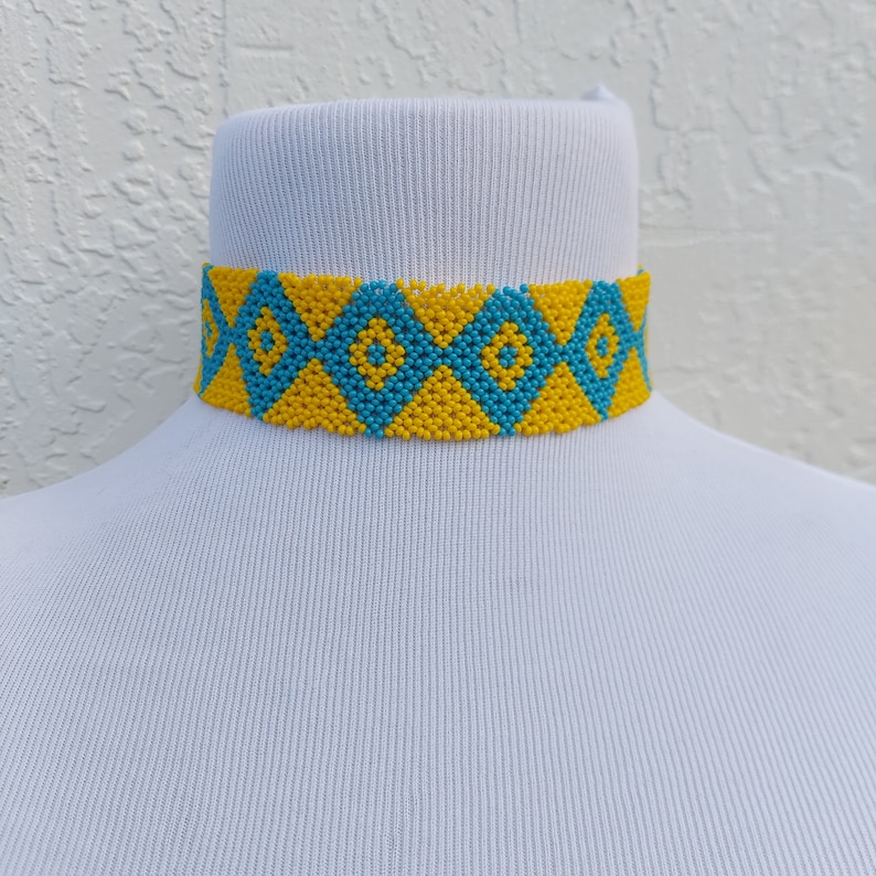May include: A beaded choker necklace in yellow and turquoise blue. The necklace features a repeating diamond pattern, with each diamond outlined in yellow and filled with turquoise beads. The necklace is displayed on a white mannequin.