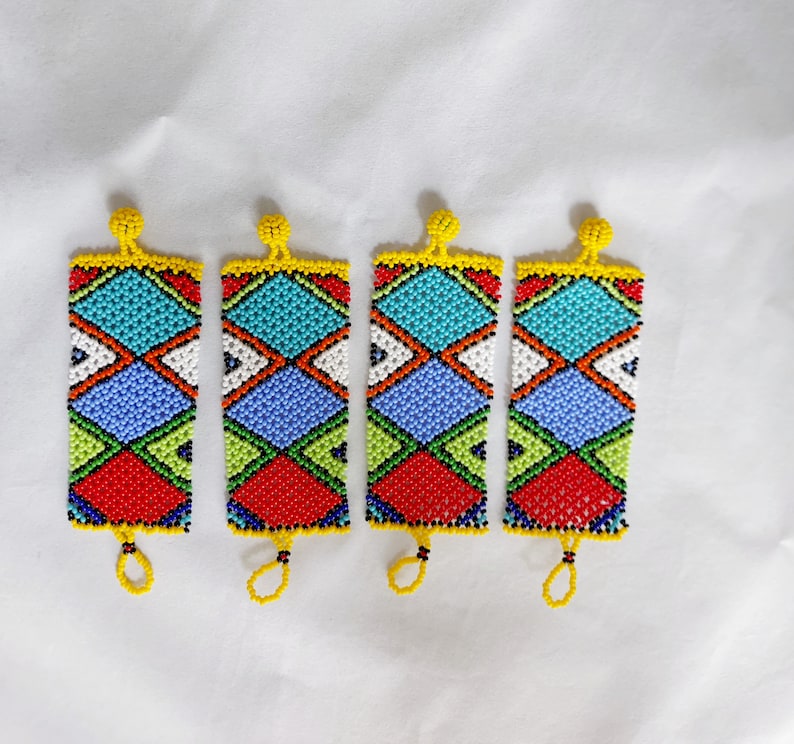 May include: Four beaded rectangular ornaments with a geometric pattern. The ornaments feature a vibrant mix of colors, including red, blue, green, white, and yellow. Each has a loop at the bottom and a beaded top.