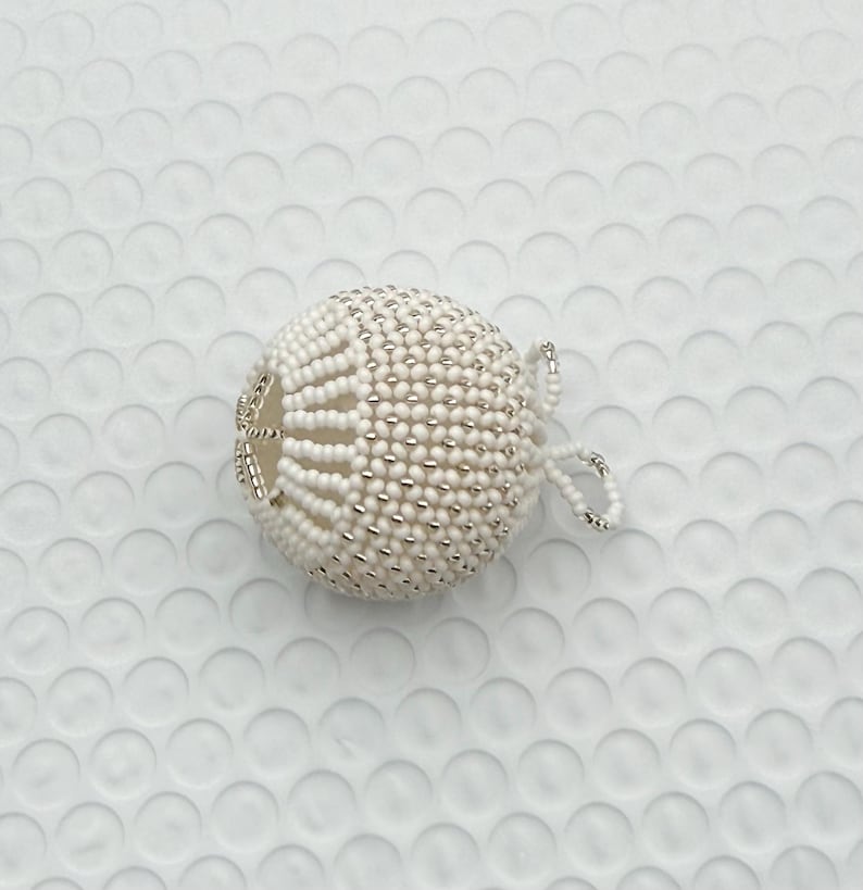 May include: A white beaded sphere ornament with a loop for hanging. The ornament is covered in small white beads with silver-colored accents. The sphere has a decorative open design on one side.