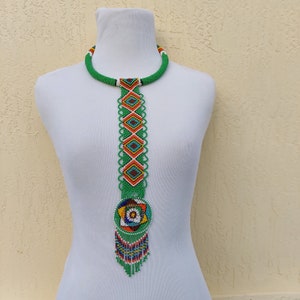 Puede incluir: Un collar de cuentas verde vibrante con un colgante circular y flecos colgantes. El collar presenta un diseño geométrico con detalles en naranja, amarillo y azul. El collar se exhibe en un maniquí blanco.