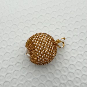 May include: A gold and white beaded ornament with a loop for hanging. The ornament is spherical, with a pattern of gold beads and white beads. The loop is made of gold and white beads.