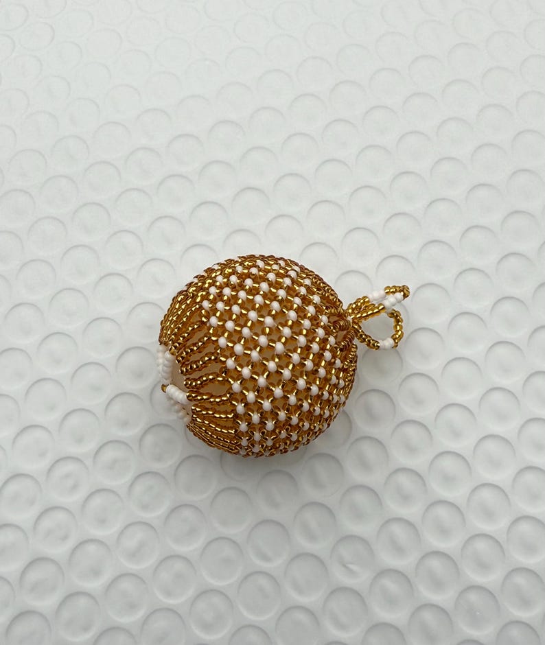 May include: A gold and white beaded ornament with a loop for hanging. The ornament is spherical, with a pattern of gold beads and white beads. The loop is made of gold and white beads.