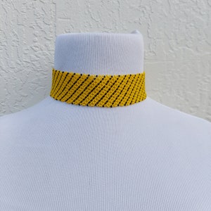 May include: A yellow beaded choker necklace with black diagonal stripes. The necklace is displayed on a white mannequin bust. The choker is a bold accessory, perfect for adding a pop of color to any outfit.