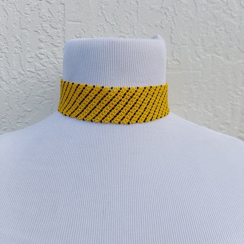 May include: A yellow beaded choker necklace with black diagonal stripes. The necklace is displayed on a white mannequin bust. The choker is a bold accessory, perfect for adding a pop of color to any outfit.