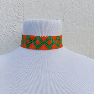 May include: A beaded choker necklace with a geometric pattern. The necklace features a repeating diamond design in orange and green. The choker is displayed on a white mannequin.