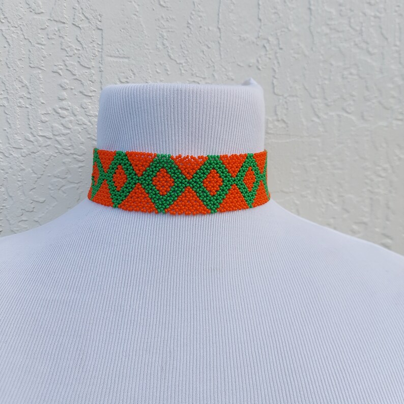 May include: A beaded choker necklace with a geometric pattern. The necklace features a repeating diamond design in orange and green. The choker is displayed on a white mannequin.