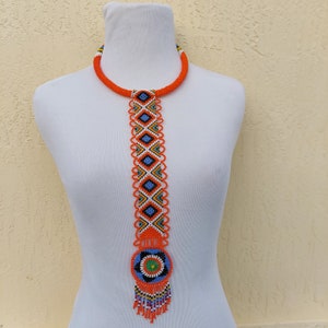 Puede incluir: Un collar de cuentas naranja con un colgante largo y vertical. El colgante presenta un patrón geométrico en azul, blanco y naranja, con un diseño circular en la parte inferior. El collar se muestra en un maniquí blanco.