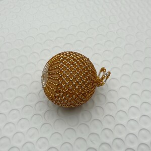 May include: A gold beaded sphere ornament with a silver wire loop for hanging. The sphere is made of small, interconnected gold beads, creating a textured surface. The ornament is likely for holiday or decorative use.