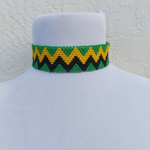 May include: A beaded choker necklace with a zig-zag pattern in green, yellow, and black. The necklace is displayed on a white mannequin head. The choker is approximately 1 inch wide.