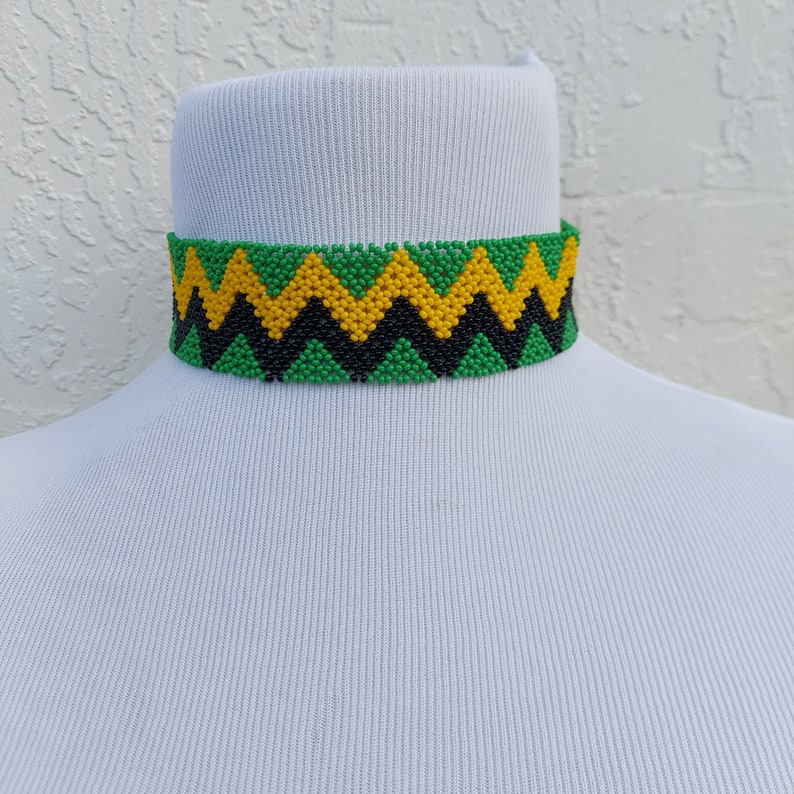 May include: A beaded choker necklace with a zig-zag pattern in green, yellow, and black. The necklace is displayed on a white mannequin head. The choker is approximately 1 inch wide.