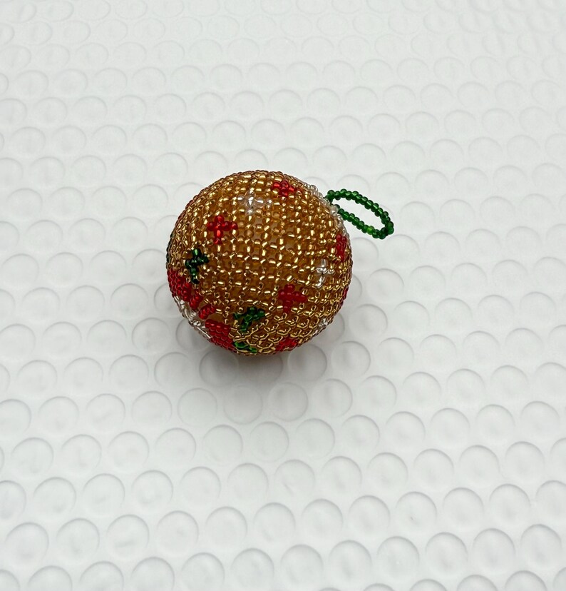 May include: A beaded Christmas ornament. The ornament is a sphere, primarily gold with red and green accents. It has a green loop for hanging.