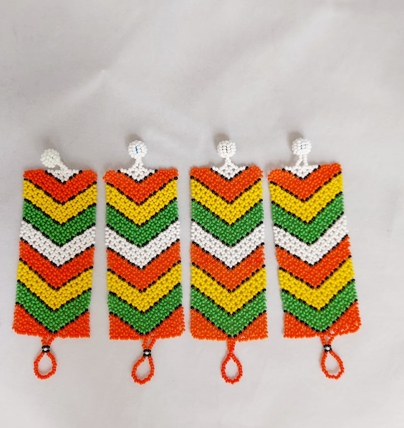 May include: Four beaded earrings with a rectangular shape and a chevron pattern. The earrings feature a color scheme of orange, yellow, green, white, and black beads. Each earring has a small loop at the bottom and a white bead at the top.