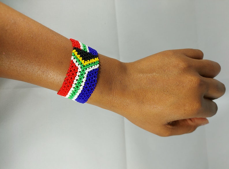May include: A beaded bracelet displaying the South African flag design. The bracelet features a pattern of red, white, green, blue, yellow, and black beads. This handmade accessory is a colourful representation of the South African flag.
