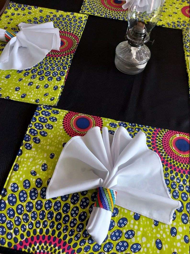 May include: A table setting featuring a vibrant green and patterned placemat with a white napkin held by a beaded napkin ring. The placemat has a colorful design with blue, pink, and yellow accents. A clear glass oil lamp sits on a black placemat.
