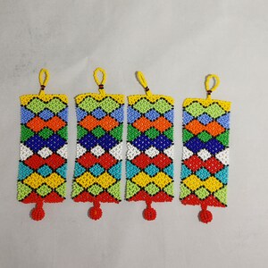 May include: Four beaded rectangular ornaments with a geometric pattern in red, orange, yellow, green, blue, and white. Each ornament has a yellow loop at the top and a red beaded tassel at the bottom. The ornaments are displayed against a light gray background.