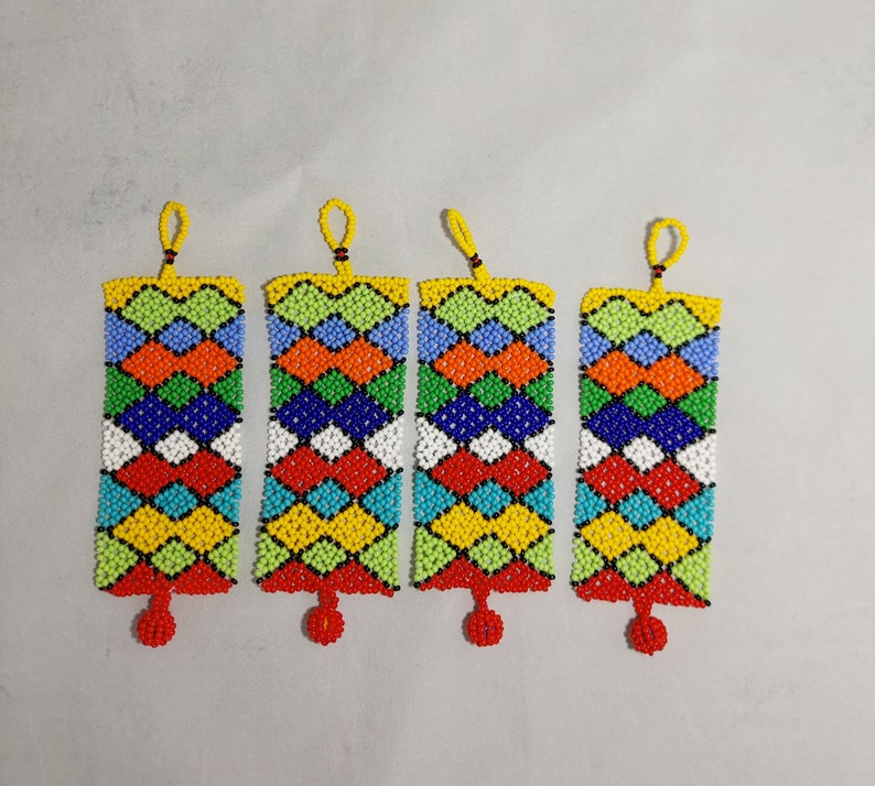 May include: Four beaded rectangular ornaments with a geometric pattern in red, orange, yellow, green, blue, and white. Each ornament has a yellow loop at the top and a red beaded tassel at the bottom. The ornaments are displayed against a light gray background.