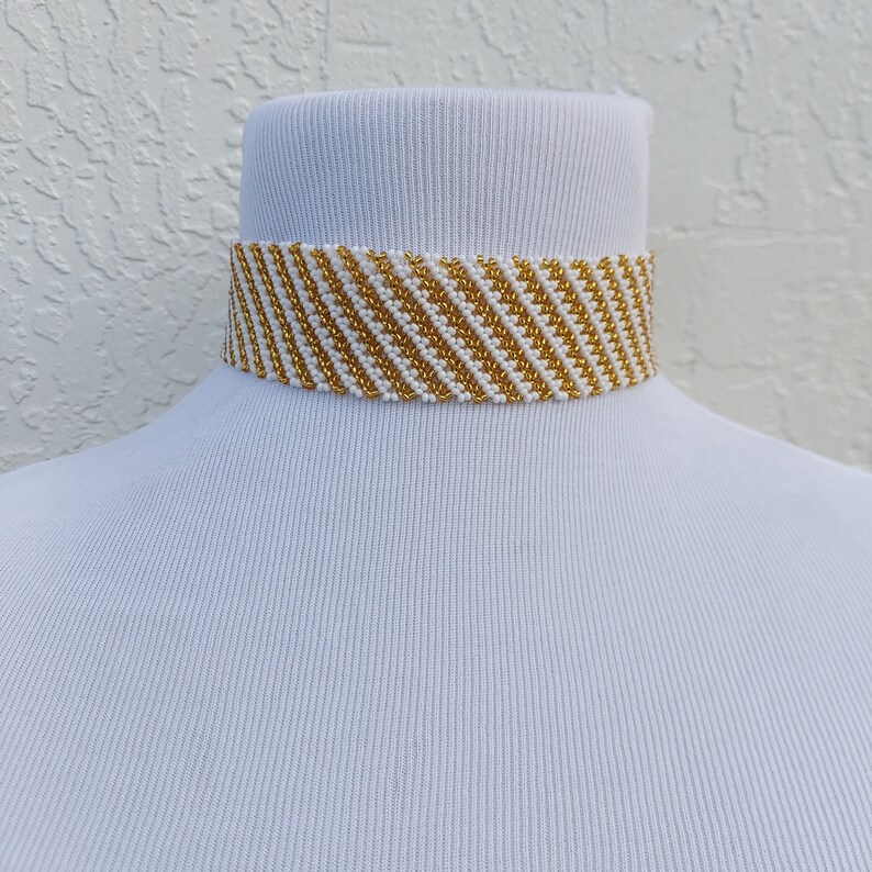 May include: A beaded choker necklace with a diagonal striped pattern. The necklace features alternating rows of gold and white beads. The choker is displayed on a white mannequin neck against a textured white background.