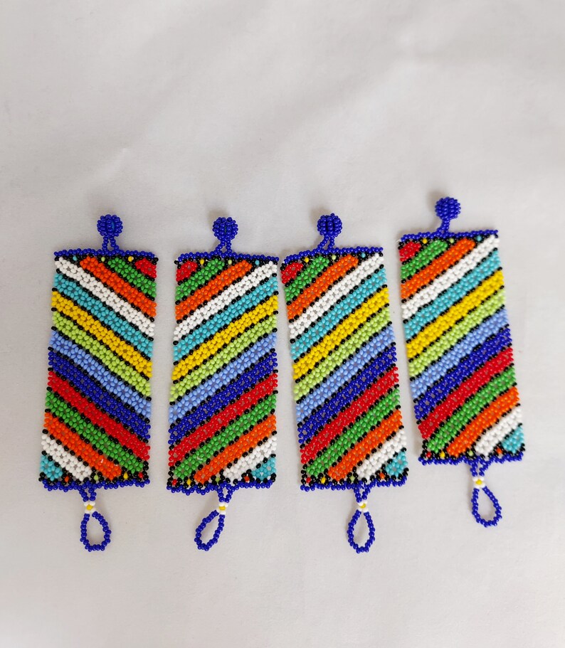 May include: Four beaded rectangular ornaments with a vibrant, diagonal striped pattern in red, orange, yellow, green, blue, and white. Each ornament has a blue loop at the bottom and a blue bead at the top. The background is plain white.