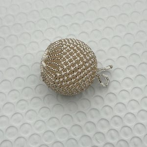 May include: A beaded sphere ornament with a loop for hanging. The ornament is made of small, silver-colored beads woven around a cream-colored core. The sphere is approximately 2 inches in diameter, with a delicate, textured appearance.