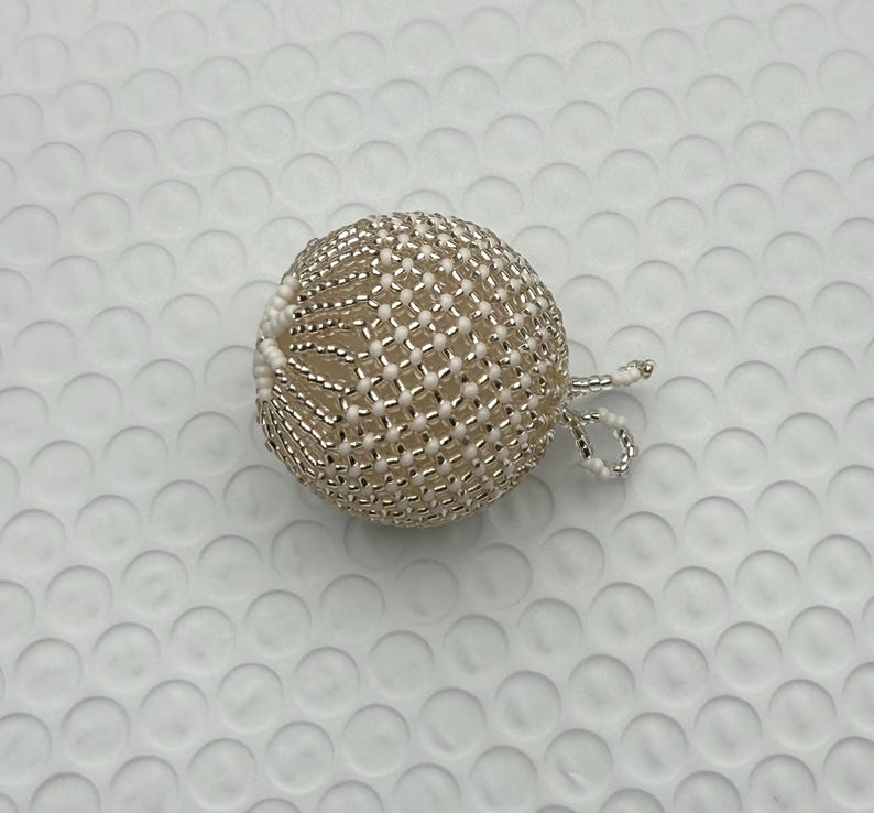 May include: A beaded sphere ornament with a loop for hanging. The ornament is made of small, silver-colored beads woven around a cream-colored core. The sphere is approximately 2 inches in diameter, with a delicate, textured appearance.