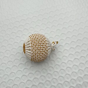 May include: A white beaded sphere with gold accents. The sphere is intricately crafted with small white beads and interspersed with gold beads, creating a textured surface. A small loop of gold beads is attached.