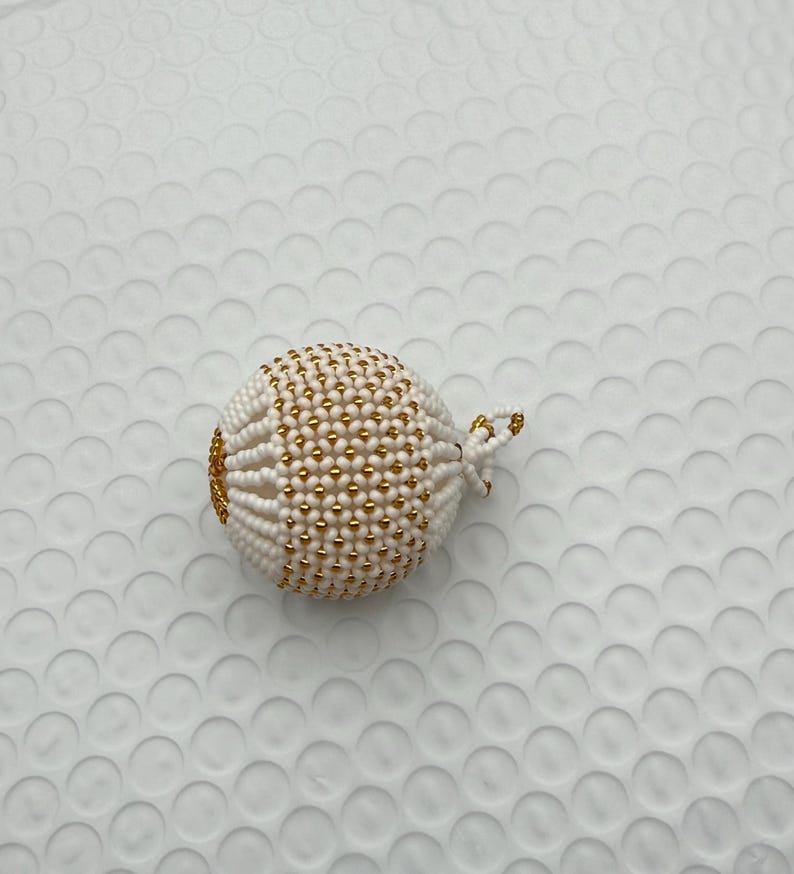 May include: A white beaded sphere with gold accents. The sphere is intricately crafted with small white beads and interspersed with gold beads, creating a textured surface. A small loop of gold beads is attached.