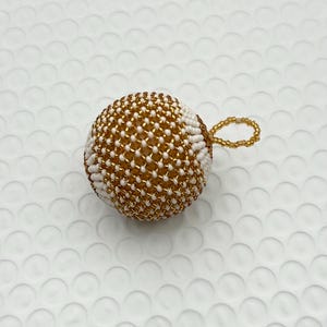 May include: A beaded sphere ornament with a gold and white pattern. The sphere has a loop for hanging. The ornament is likely handmade and could be used as a Christmas tree decoration or a decorative accent.