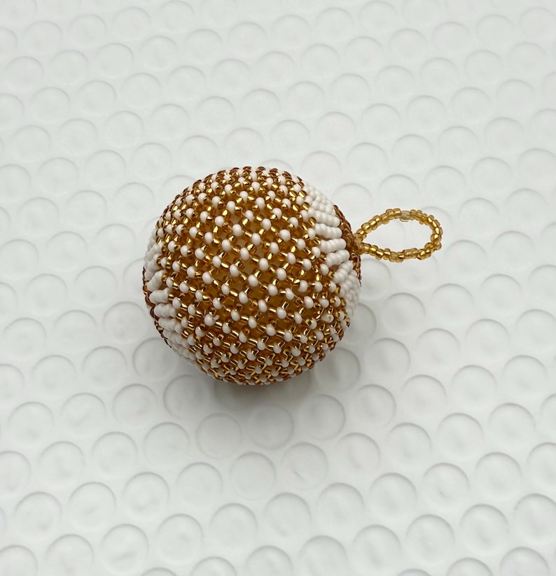 May include: A beaded sphere ornament with a gold and white pattern. The sphere has a loop for hanging. The ornament is likely handmade and could be used as a Christmas tree decoration or a decorative accent.