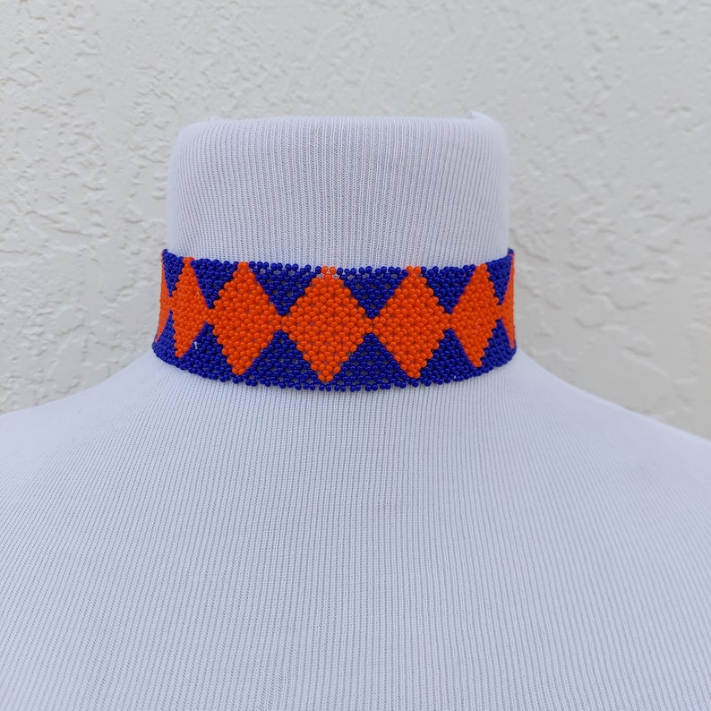 May include: A beaded choker necklace with a geometric pattern. The choker features a repeating diamond design in orange and blue. The necklace is displayed on a white mannequin neck.