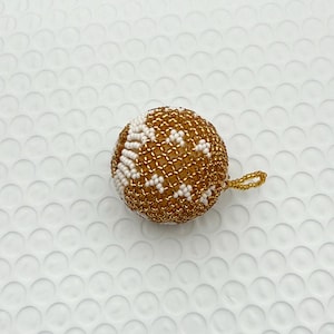 May include: A round, beaded ornament with a gold and white pattern. The ornament has a small gold loop for hanging. The beads are tightly woven together, creating a textured surface. The ornament is likely for holiday or seasonal decor.