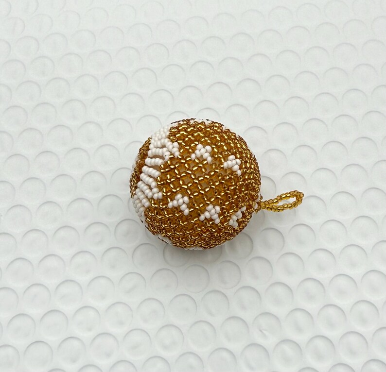 May include: A round, beaded ornament with a gold and white pattern. The ornament has a small gold loop for hanging. The beads are tightly woven together, creating a textured surface. The ornament is likely for holiday or seasonal decor.