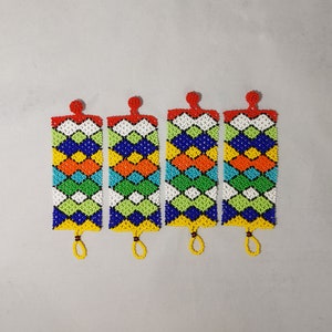 May include: Four beaded rectangular ornaments with a geometric pattern in blue, orange, yellow, green, and white. Each has a red beaded top and a yellow beaded loop at the bottom. The ornaments are displayed on a light gray background.