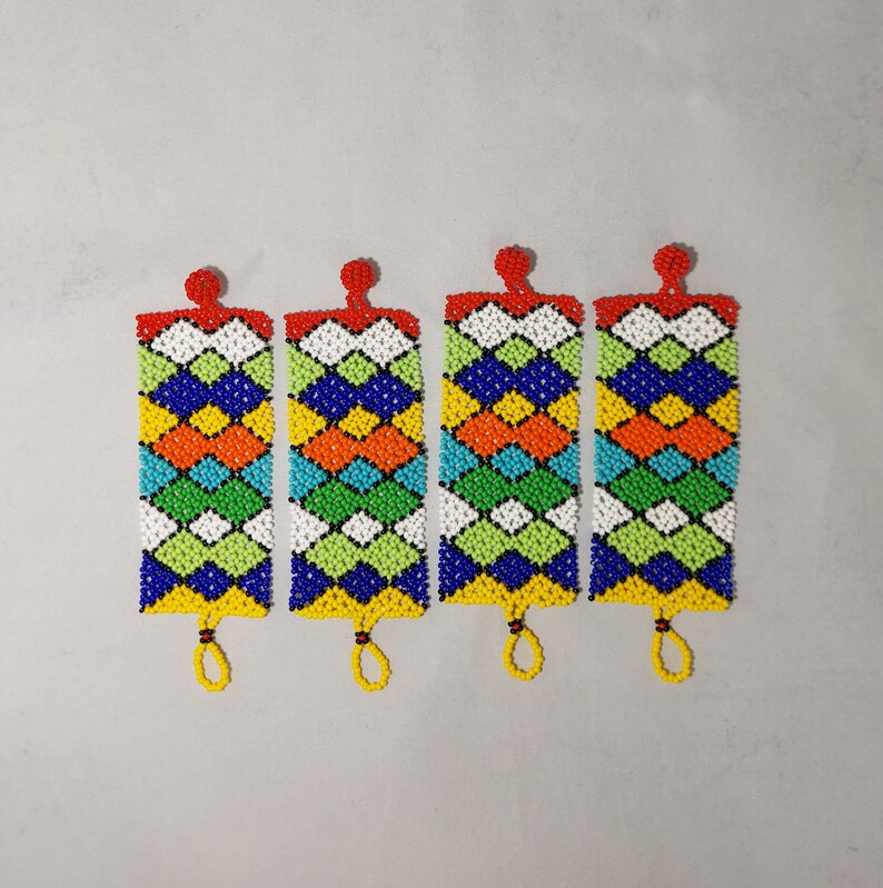 May include: Four beaded rectangular ornaments with a geometric pattern in blue, orange, yellow, green, and white. Each has a red beaded top and a yellow beaded loop at the bottom. The ornaments are displayed on a light gray background.