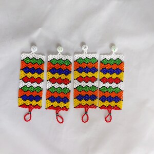 May include: Four beaded rectangular ornaments with a white background. Each ornament features a zig-zag pattern in red, orange, green, yellow, blue, and white beads. A small red loop hangs from the bottom of each ornament. The top has a white beaded ball.