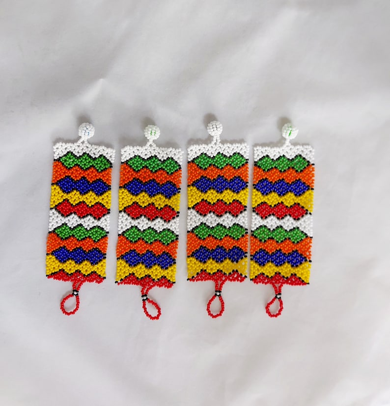 May include: Four beaded rectangular ornaments with a white background. Each ornament features a zig-zag pattern in red, orange, green, yellow, blue, and white beads. A small red loop hangs from the bottom of each ornament. The top has a white beaded ball.