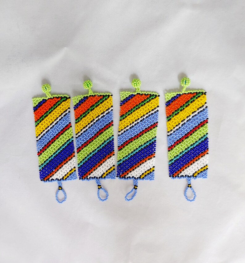 May include: Four beaded bookmarks with a colorful striped pattern. The bookmarks feature a variety of colors, including red, orange, yellow, green, blue, and white. Each bookmark has a small loop at the bottom and a green beaded top.