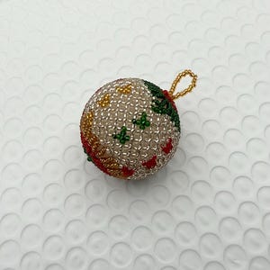 Puede incluir: Un adorno navideño con cuentas con un lazo para colgar. El adorno es una esfera cubierta de pequeñas cuentas transparentes. Tiene detalles de cuentas rojas, verdes y amarillas. El lazo está hecho de cuentas doradas.