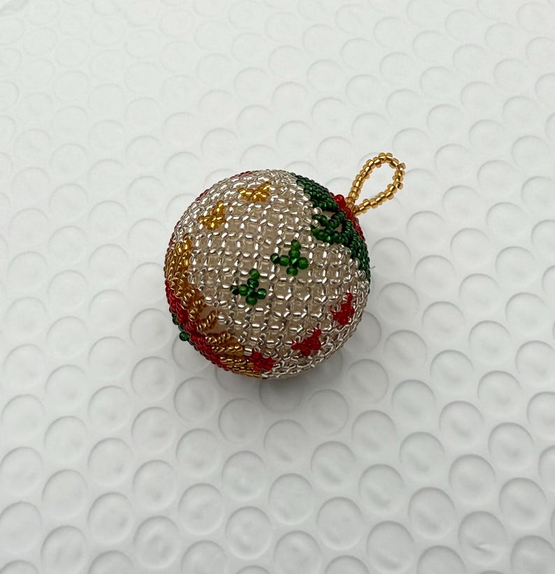 May include: A beaded Christmas ornament with a loop for hanging. The ornament is a sphere covered in small, clear beads. It has red, green, and yellow beaded accents. The loop is made of gold-colored beads.