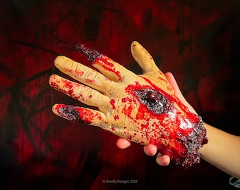 May include: A prosthetic hand with realistic skin tones, featuring a large, open wound and severed fingertips. The hand is covered in fake blood, with a dark red background. The image is labeled "Deadly Designs 2022".