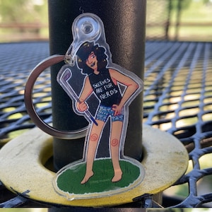 May include: A clear acrylic keychain featuring a cartoon character with long black hair, wearing a black tank top with the text "Sleeves are for Nerds" and blue and white patterned shorts, holding a golf club. The character is standing on a green patch of grass.
