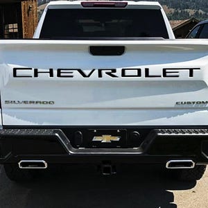 Puede incluir: Vista trasera de una camioneta Chevrolet Silverado blanca. La palabra "CHEVROLET" está en la puerta trasera en letras negras. Debajo, también son visibles "SILVERADO" y "CUSTOM". La camioneta tiene un parachoques negro y tubos de escape.