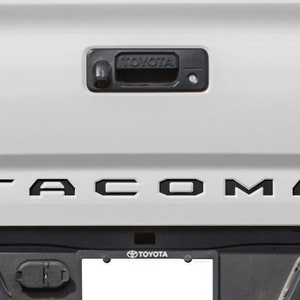 May include: A white pickup truck tailgate with a black "TACOMA" lettering across the top and a black "TOYOTA" emblem on the lower right side.