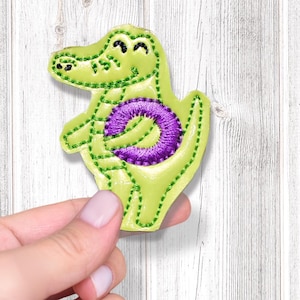 May include: A lime green alligator applique with a purple belly. The alligator has a smiling face and is made of fabric with stitched details. The applique is held in front of a light wood-grain background.