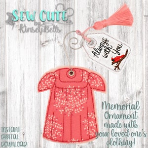May include: A pink fabric ornament shaped like a dress with a white floral pattern. The ornament is attached to a metal hanger with a tag that reads "Always with You" and features a red cardinal.